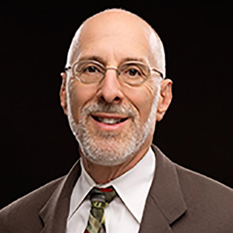 Sokoloff is pictured against a black backdrop wearing glasses and a brown suit jacket