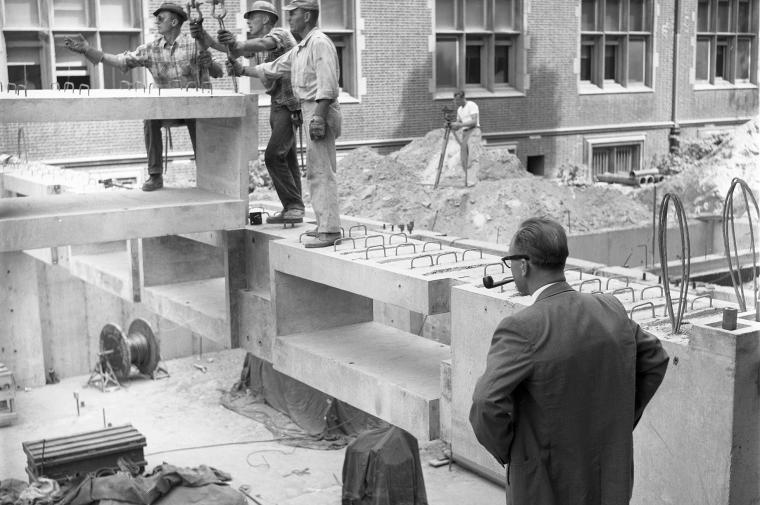 B/W photo of men working at construction site with other man watching them while smoking a pipe