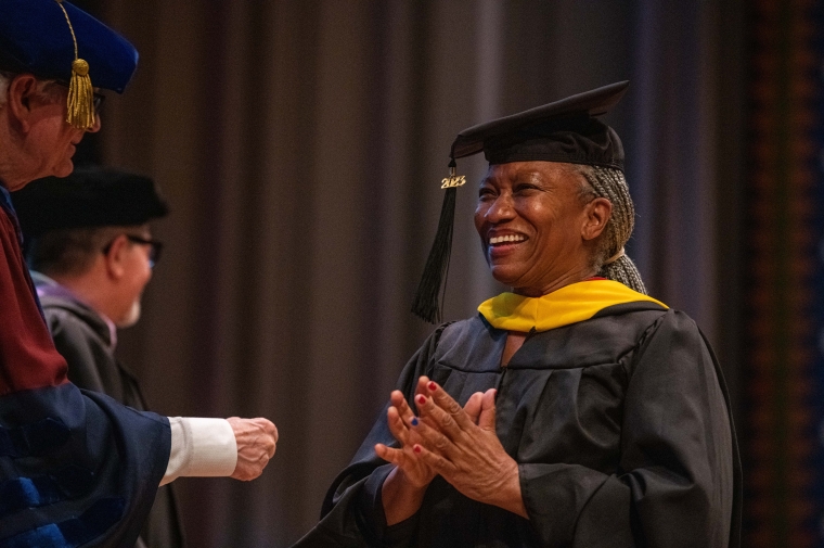 A woman in cap and gown holds hands to her chest in appreciation