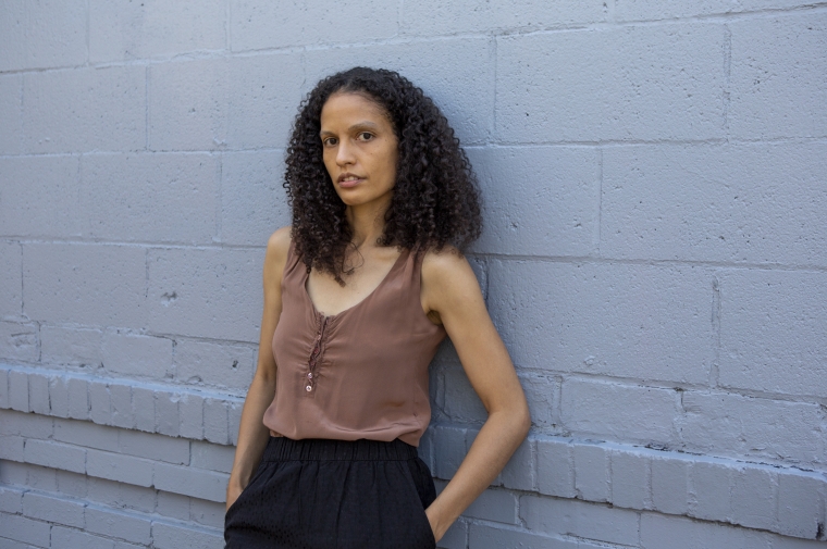 Vaughn leaning back on a blue painted brick wall, hands in her pockets