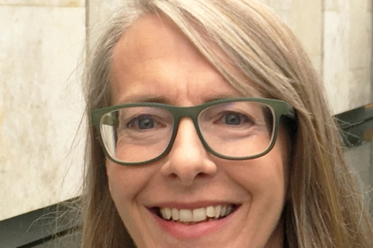 Neises looks into the camera, smiling, wearing dark rimmed glasses and against a black and white background