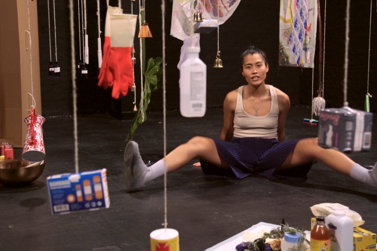 Performer sitting on ground in black box, with hanging objects in the foreground to background