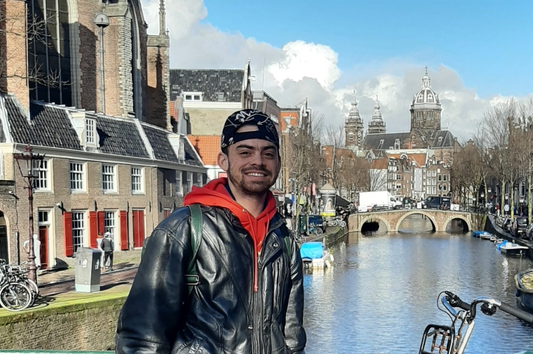 A photograph of Geo Wyex standing on a bridge in front of a river surrounded by historic houses