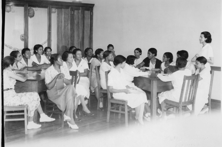 B&W photo of group of women having meeting around several tables