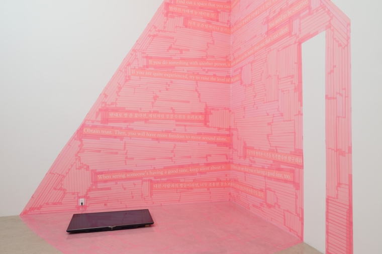 Photograph of corner installation of strips of pink tape in geometric shapes with white text in Korean and English