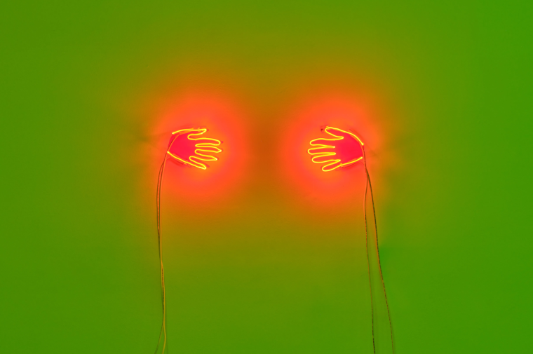 Two orange neon hands with fingers facing towards each other mounted on a green wall over a soil floor
