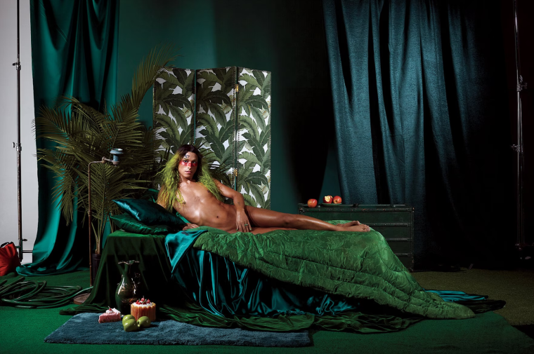 Photography of the artist, Puppies Puppies (Jade Guanaro Kuriki-Olivo), laying on a green covered bed in a classic nude half-reclining pose, looking at the viewer