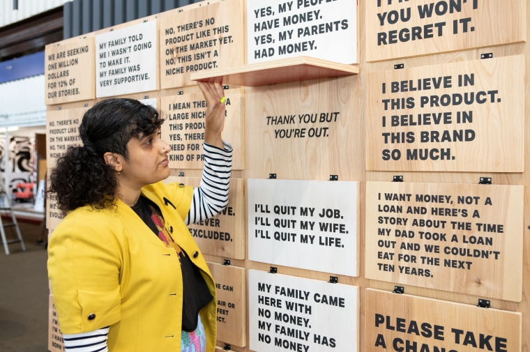 A woman in a yellow blazer holds open a wooden flap with text inscribed on its face and behind