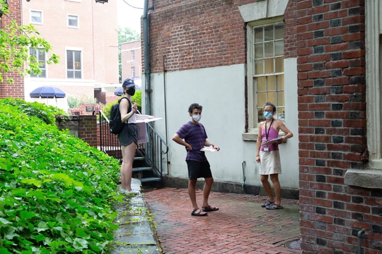 Three people in face masks stand with clipboards outdoors next to a brick building.