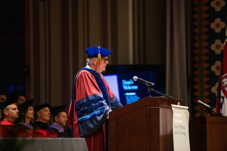 Man in burgundy and blue graduation robe at a podium