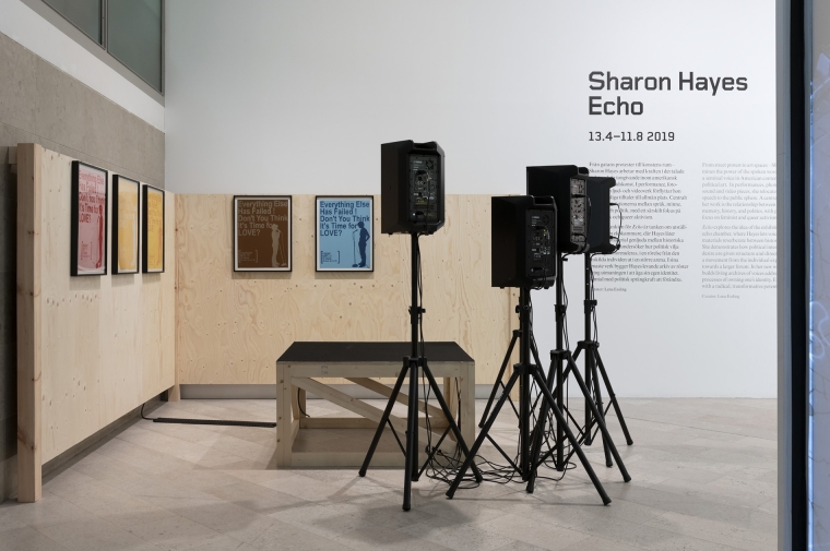 Four speakers on stands and framed posters next to wall in gallery