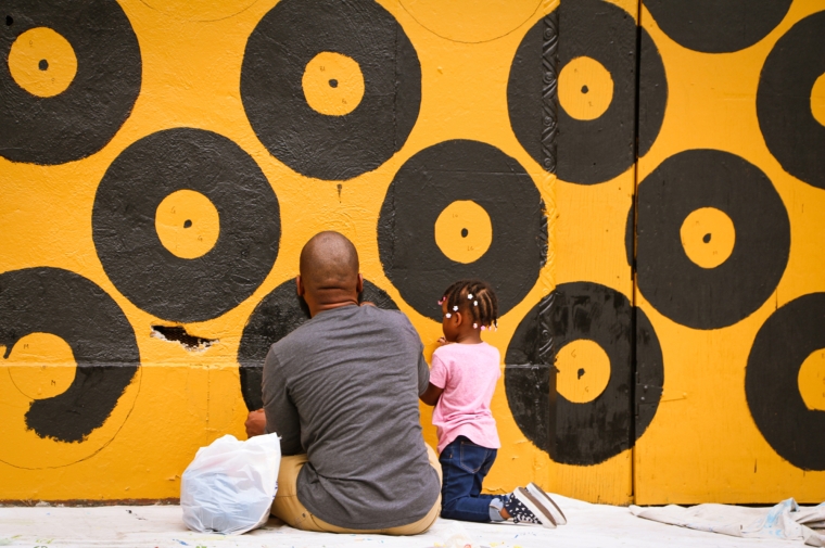 An African American man and young girl seated in front of a black and gold mural