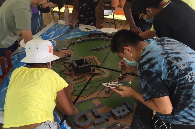 a group of young people huddle around a green architectural model