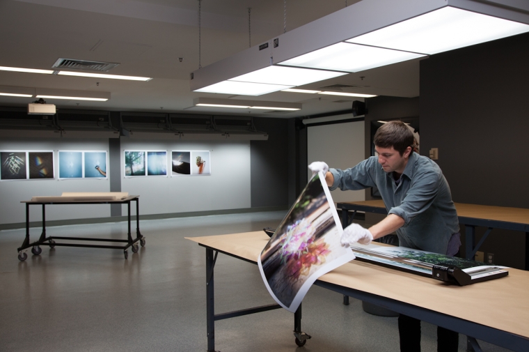 Student prepares to trim a large photograph in a softly lit studio. Large prints of photographs hang on the wall in the back