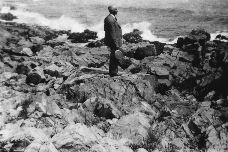 A middle aged man in a business suit stands on a rocky shore, looking at the ocean