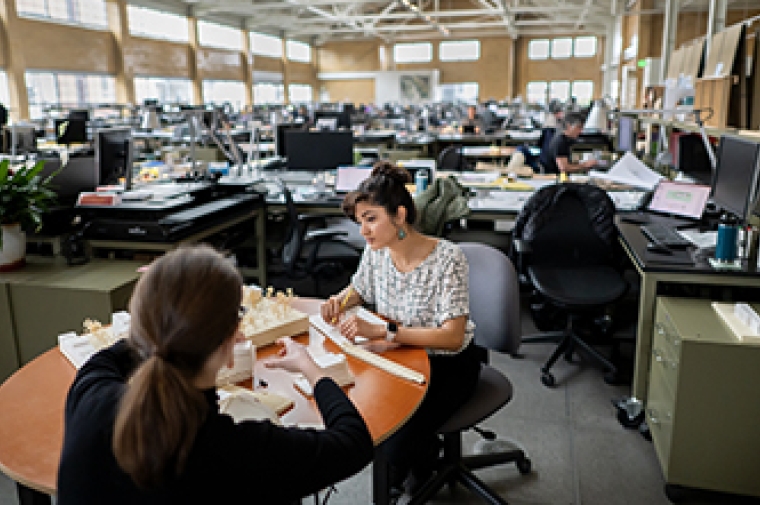 Two women seated at table work on wooden architectural model