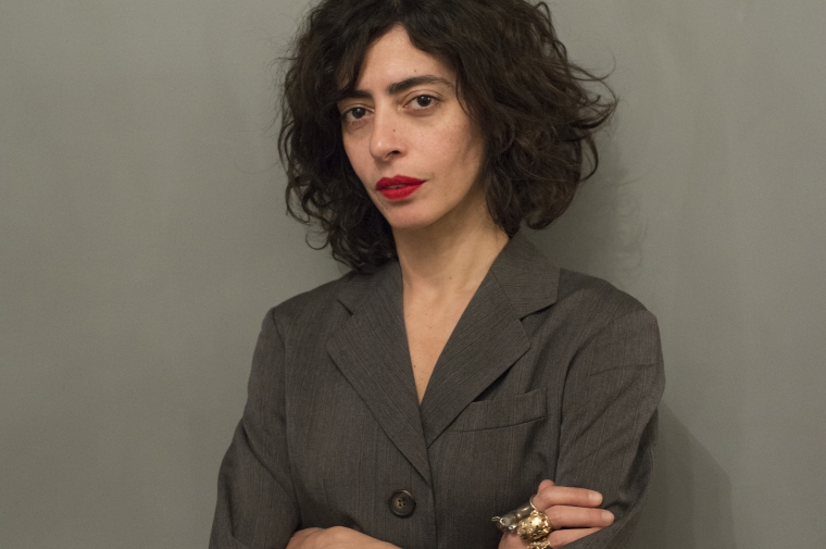 headshot of maria with her arms folded, wearing a gray blazer on a gray background