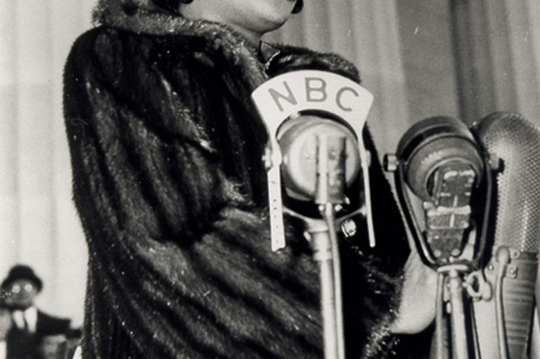 A young Black woman in a fur coat sings at a microphone