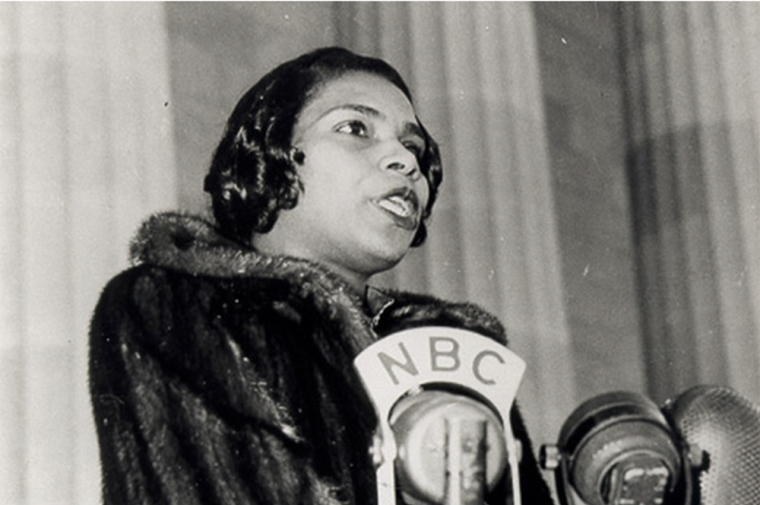 A young Black woman in a fur coat sings at a microphone