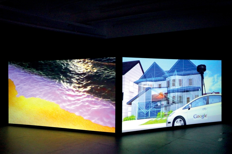 Photograph of gallery installation of two large screens, one displaying a brightly colored beach, the other depicting a google car in front of a suburban home