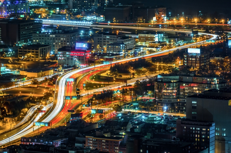 Car headlights and taillights on elevated highway weave between office and apartmentbuildings 