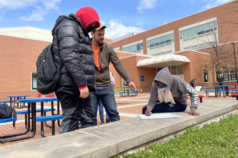 A man gestures to a drawing with two young people outside a brick building