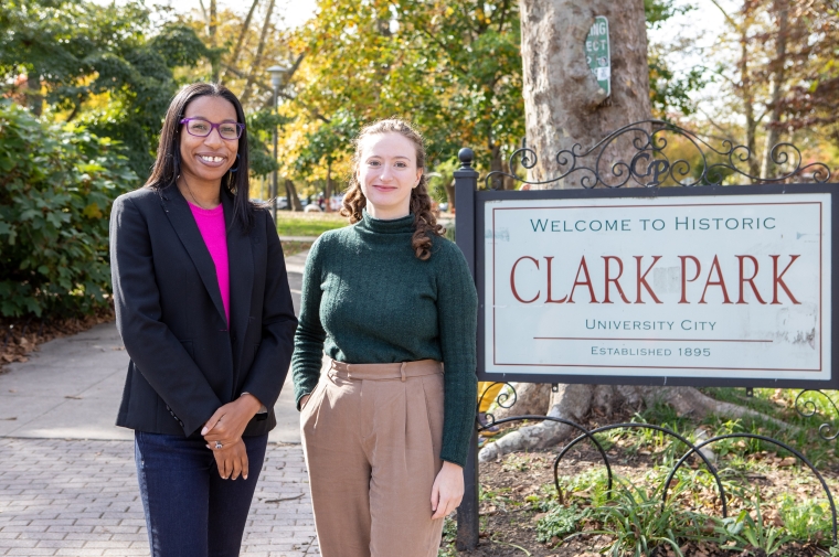 A woman in a dark suit and a woman in a green sweater stand outdoors next to a sign for "Clark Pakr"