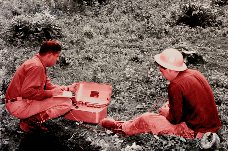 Black and white background of landmine exploding with two men colored in red tones in the front observing