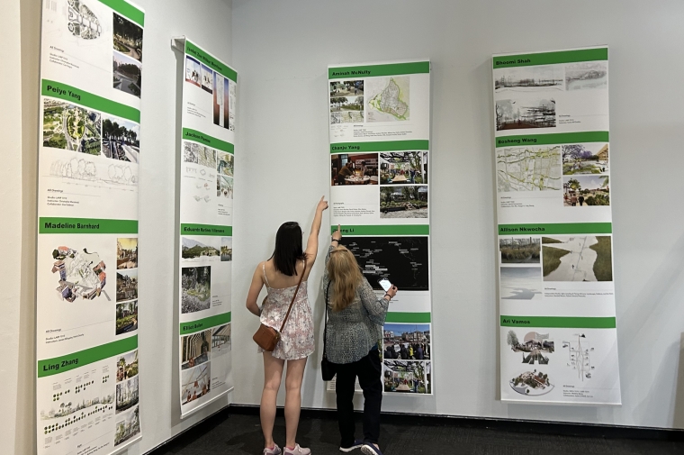 Two women gesture towards a printed rendering hung in a white gallery