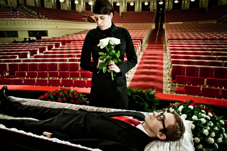 Woman holding flowers standing over man lying in coffin