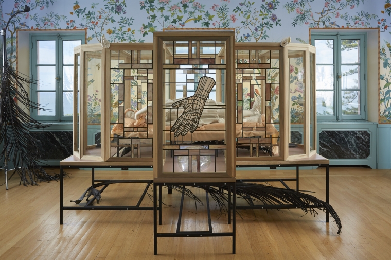 installation view of glass box with body inside, stained glass hand obstructing the view, in an ornate room