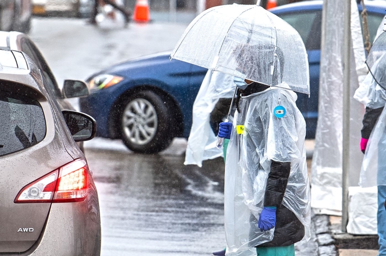 A healthcare worker in full PPE stands next to a car in the rain