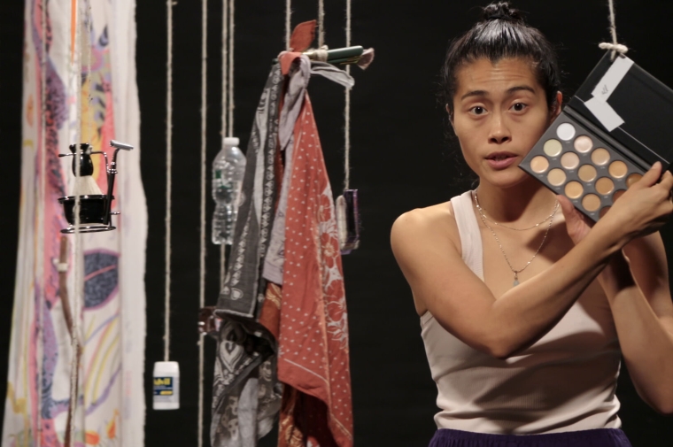 Video still from "One Big Bag" which shows a woman displaying a makeup palette with various skin tones and other items hung next to her: a shaving kit, hairbrush, water bottle, container of Advil, bandanas, and others