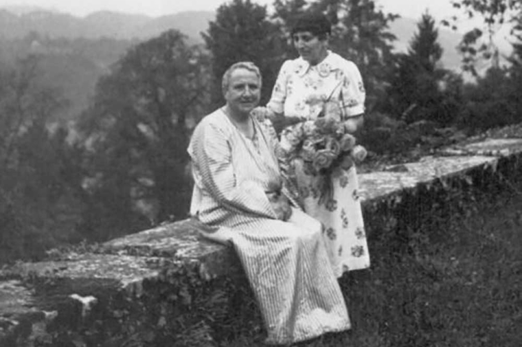 Two women pose outdoors, one of them is seated on a low wall