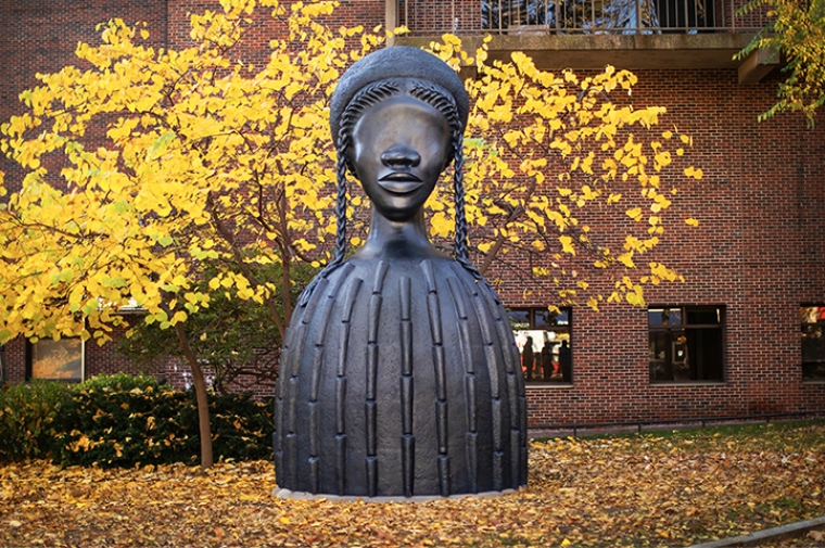 Large bronze sculpture of a woman seen against autumn trees