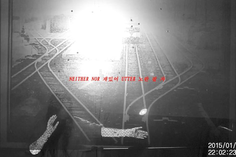 black and white video still of the artist behind a mesh screen with railroad tracks and text projected onto it