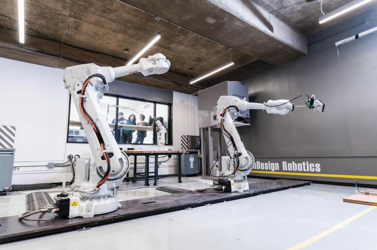 Two large robotic arms in a studio with students viewing them through a window to an adjacent room