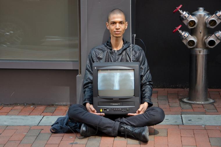 Wilson sitting cross-legged on a city street, staring directly at camera with vintage TV in their lap