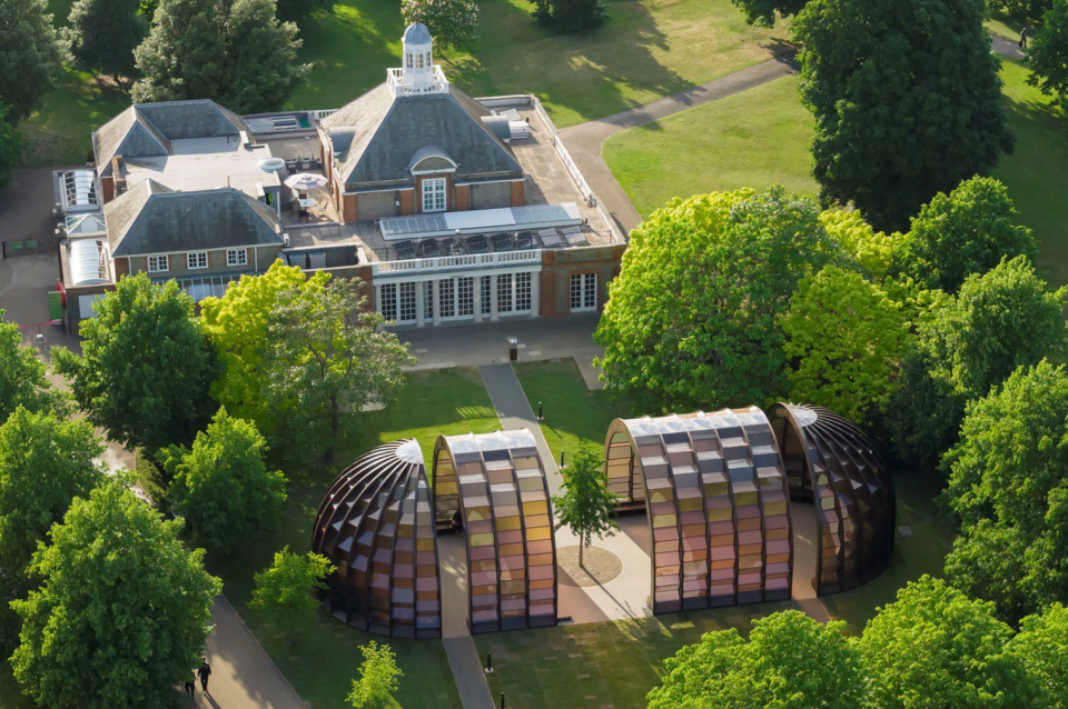 A capsule-like pavilion adjacent to a squat 19th century building in a green park 