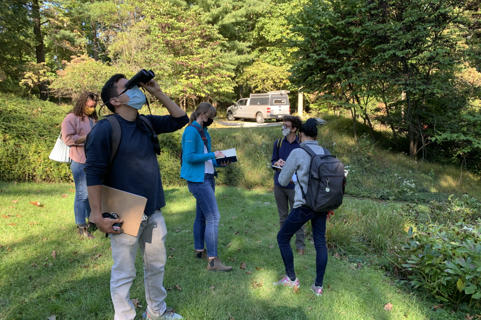 Students stand in shady grass area, one of them holding binoculars