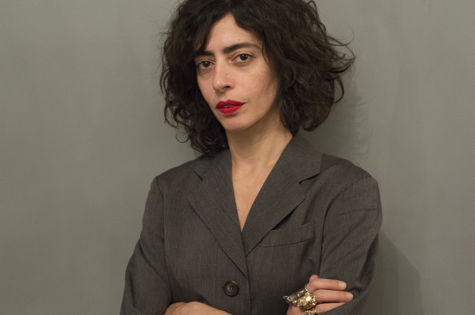 headshot of maria with her arms folded, wearing a gray blazer on a gray background