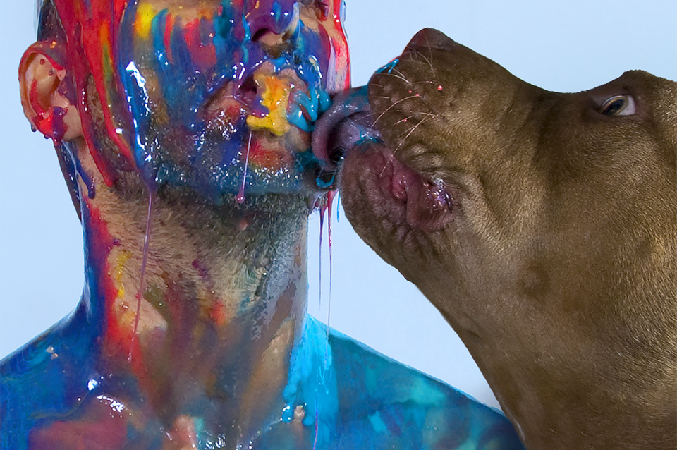 Self-portrait of the artist covered in brightly colored viscous liquid with a dog licking his face 