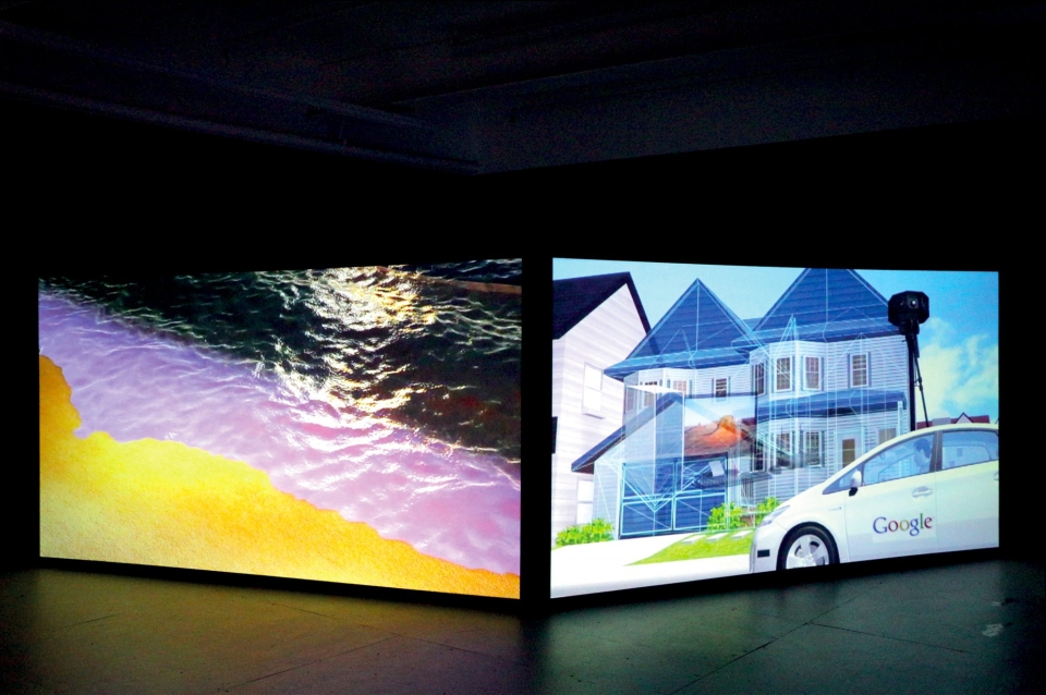 Photograph of gallery installation of two large screens, one displaying a brightly colored beach, the other depicting a google car in front of a suburban home