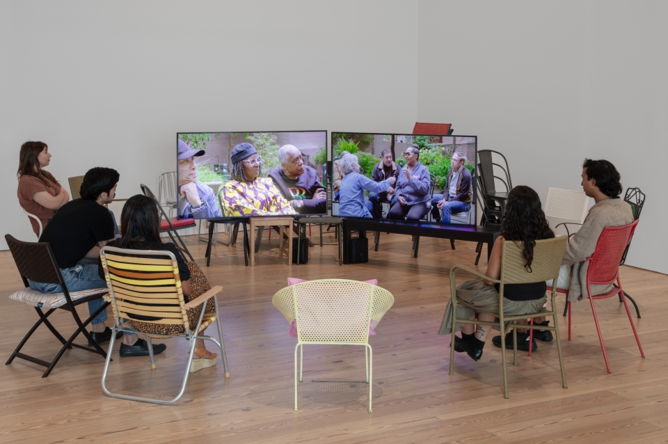 five people sitting in outdoor chairs assembled in a circle watching two video monitors with images of a group of people gathered together being interviewed.