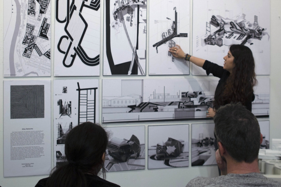 a woman pointing to large architectural drawings hung on the wall.
