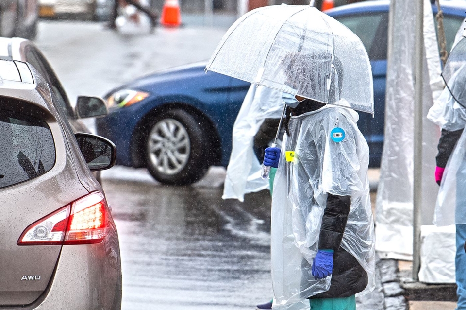 A healthcare worker in full PPE stands next to a car in the rain