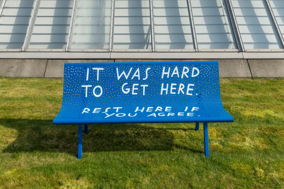 Photograph of a blue bench outside on a lawn with the text "IT WAS HARD TO GET HERE. REST HERE IF YOU AGREEE".