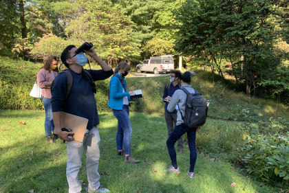 Students stand in shady grass area, one of them holding binoculars 