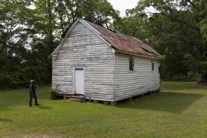 Weathered single-story white wood-sided structure with red roof in a wooded setting