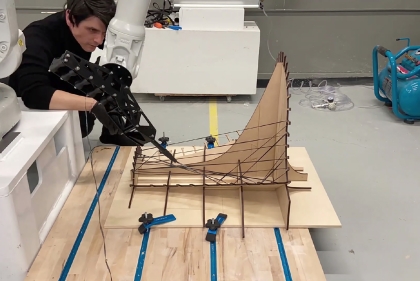 A man watches a robot arm weave black fibers on a wood frame